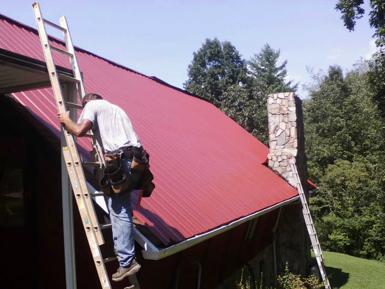 Roofer on a ladder beside a red metal roof and stone chimney — Soffit & Fascia Repair by Roofing Brothers in Red Lake Falls